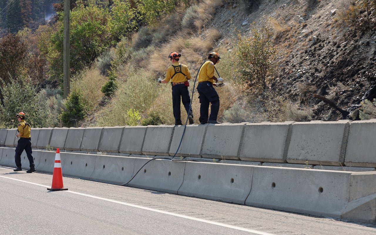 default-1589 Firefighters in yellow gear near a highway, using hoses, with charred ground visible.